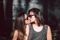 Zarte Liebhaberinnen in lässigem Outfit und Sonnenbrille küssen sich, während sie an einem sonnigen Sommertag draußen auf verschwommenem Straßenhintergrund spazieren — Stockfoto