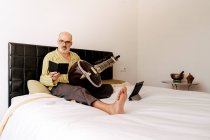 Älterer Mann sitzt mit Sitar im Bett und macht sich Notizen in Notizbuch, während er Online-Unterricht auf dem Tablet schaut — Stockfoto