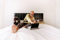 Літній чоловік сидить на ліжку з ситаром і робить нотатки в блокноті під час перегляду онлайн-уроку на планшеті — стокове фото