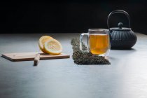 Bevanda aromatica in tazza di vetro e teiera disposti con limoni e cumuli di foglie di tè essiccate sul tavolo su sfondo nero — Foto stock