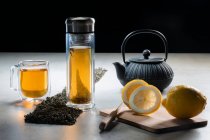 Bevanda aromatica in tazza di vetro e teiera disposti con limoni e cumuli di foglie di tè essiccate sul tavolo su sfondo nero — Foto stock