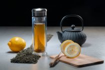 Bebida aromática em caneca de vidro e bule dispostas com limões e montes de folhas de chá secas na mesa sobre fundo preto — Fotografia de Stock