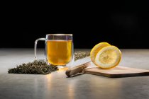 Bebida aromática em caneca de vidro arranjada com limões e montes de folhas de chá secas na mesa sobre fundo preto — Fotografia de Stock