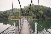 Женщина-путешественница в пальто фотографируется с фотокамерой, стоя на деревянном подвесном мосту с металлическим забором над озером возле зеленого леса — стоковое фото