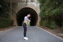 Vista lateral de delgada hembra asiática joven en leggings de pie en la carretera de asfalto frente al túnel cerca de la pared cubierta de plantas verdes y mirando a la cámara - foto de stock