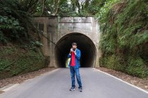 Чоловічий турист у червоній сорочці та джинсовій куртці фотографується на цифровій фотокамері, стоячи на асфальтній дорозі біля зеленого кріплення та тунелю вдень — стокове фото