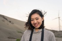 Calm young Asian female in casual wear touching hair and looking at camera while standing near seacoast under cloudy sky on windy day — Stock Photo