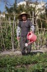 Full body middle aged Asian man in traditional oriental straw hat looking at camera and using watering pot while pouring green plants growing in garden in Taiwan — Stock Photo