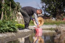 Side view of focused Asian man in dirty clothes with oriental straw hat filling plastic watering pot for pouring plants while standing beside narrow creek on farm in Taiwan — Stock Photo