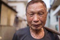 Retrato de anciano asiático jardinero en ropa casual sonriendo a la cámara mientras está de pie en la calle con azada sobre el hombro en el asentamiento en Taiwán - foto de stock