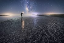 Back view of anonymous man standing and holding torch on empty road among calm water and reaching out to star under colorful nigh sky with milky way on background — Stock Photo