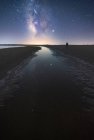 Vista posterior del hombre anónimo de pie en el camino vacío entre el agua tranquila y llegar a la estrella bajo el colorido cielo cercano con la vía láctea en el fondo - foto de stock