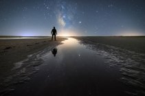 Back view of anonymous man standing on empty road among calm water and reaching out to star under colorful nigh sky with milky way on background — Stock Photo