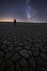 Silhouette of unrecognizable man standing on dry cracked surface of ground reaching out to a colorful night starry sky on horizon — Stock Photo