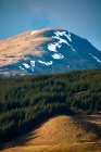 Живописный вид горного склона покрытого снегом среди зеленых холмов с лесом против облачного неба в весенний день в Шотландском нагорье — стоковое фото