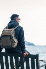 Back view of male hiker in warm jacket with backpack sitting on wooden bench near sea and enjoying marine scenery with rocky coast during travel in Scotland — Stock Photo