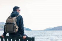 Back view of male hiker in warm jacket with backpack sitting on wooden bench near sea and enjoying marine scenery with rocky coast during travel in Scotland — Stock Photo