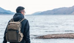 Vista trasera del excursionista masculino con chaqueta cálida con mochila sentada en un banco de madera cerca del mar y disfrutando de paisajes marinos con costa rocosa durante el viaje en Escocia - foto de stock