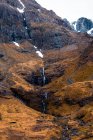 Pendiente rocosa áspera de la montaña con arbustos sin hojas y cascada en las tierras altas escocesas - foto de stock