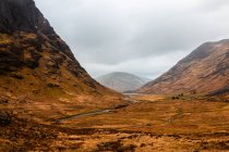 Вузька нерівна дорога проходить гористою місцевістю з сухою травою серед скелястих гір у похмурий весняний день у Шотландському нагір 