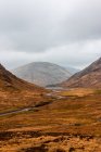 Carretera estrecha con curvas que atraviesa un terreno montañoso con hierba seca entre montañas rocosas en un día nublado de primavera en las tierras altas escocesas - foto de stock