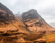 Schmale kurvenreiche Straße durch hügeliges Gelände mit trockenem Gras zwischen felsigen Bergen an bewölkten Frühlingstagen in den schottischen Highlands — Stockfoto