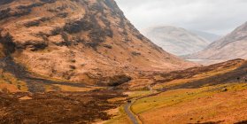 Carretera estrecha con curvas que atraviesa un terreno montañoso con hierba seca entre montañas rocosas en un día nublado de primavera en las tierras altas escocesas - foto de stock
