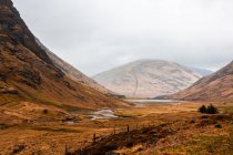 Вузька нерівна дорога проходить гористою місцевістю з сухою травою серед скелястих гір у похмурий весняний день у Шотландському нагір 