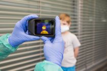 Médecin méconnaissable dans des gants de protection et uniforme à l'aide d'une caméra d'imagerie thermique pour vérifier la température de l'enfant dans la rue pendant l'épidémie de coronavirus — Photo de stock