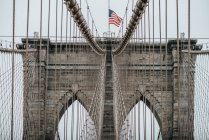 Baixo ângulo de torres de pedra com arcos pontiagudos de suspensão Ponte Brooklyn com bandeira americana em dia nublado — Fotografia de Stock
