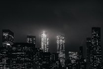 Черно-белый комплекс современных освещенных небоскребов на Манхэттене, расположенный перед спокойной рекой в ночное время — стоковое фото