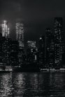 Черно-белый комплекс современных освещенных небоскребов на Манхэттене, расположенный перед спокойной рекой в ночное время — стоковое фото