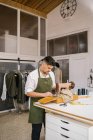 Fleißig fokussierte männliche Schneiderin in Schürze näht Outfit Details mit modernen Nähmaschine am Tisch, während die Schaffung exklusiver Kleidungskollektion in zeitgenössischen Arbeitsatelier — Stockfoto