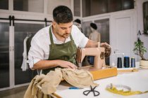 Diligent focused male tailor in apron sewing outfit details using modern sewing machine at table while creating exclusive clothes collection in contemporary work studio — Stock Photo