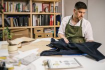 Обрізати молодого чоловіка, який працює з курткою на шиття деталей блискавки майбутнього вбрання, працюючи над ексклюзивною колекцією одягу за столом проти розмитого інтер'єру сучасного ательє — стокове фото