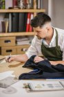 Crop young male dressmaker writing with pen on sewing pattern examining or developing details of future outfit while working on exclusive apparel collection at table against blurred interior of contemporary atelier — Stock Photo