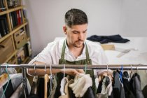 High angle of serious focused male tailor in apron checking details of apparel hanging on hanger on metal rack among other trendy bespoke clothes in modern workroom — Stock Photo