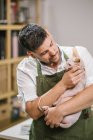 Artisan in white shirt and green apron while carry calm Sphynx cat on hands in modern studio — Stock Photo