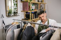High angle of serious focused male tailor in apron checking details of apparel hanging on hanger on metal rack among other trendy bespoke clothes in modern workroom — Stock Photo