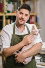 Fröhlicher unrasierter Handwerker in weißem Hemd und grüner Schürze lächelt in die Kamera, während er im modernen Studio die ruhige Sphynx-Katze auf Händen trägt — Stockfoto