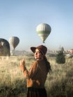 Улыбающаяся туристка в модной одежде, стоящая на зеленом поле и наслаждающаяся солнечной погодой на фоне воздушных шаров — стоковое фото