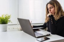 Seitenansicht einer fokussierten jungen Frau in lässigem Outfit, die mit Laptop am Tisch sitzt und Notizen im Planer macht, während sie an einem hellen Arbeitsplatz zu Hause online arbeitet — Stockfoto