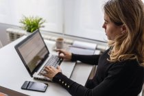 Сверху сосредоточенная молодая женщина в повседневной одежде сидит за столом с ноутбуком и работает в Интернете на легком рабочем месте дома — стоковое фото