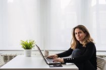 Вид сфокусированной женщины-специалиста по повседневной одежде, работающей с электронными документами на ноутбуке, сидя за столом у окна в современной комнате дома — стоковое фото