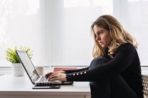 Высокоугольный вид сбоку сфокусированной женщины-специалиста по повседневной одежде, работающей с электронными документами на ноутбуке, сидя за столом у окна в современной комнате дома — стоковое фото