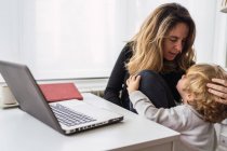 Moderne geschäftige Unternehmerin in Freizeitkleidung umarmt und küsst kleines Kind, während sie im Homeoffice an einem Projekt mit Laptop arbeitet — Stockfoto