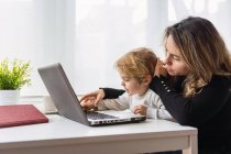 Side view of female remote employee holding curious little child on knees while sitting at table and working with laptop at home — Stock Photo