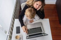 Зверху жіночий віддалений працівник тримає допитливу маленьку дитину на колінах, сидячи за столом і працюючи з ноутбуком вдома — стокове фото