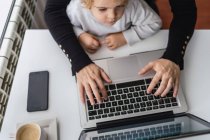 From above of crop anonymous female remote employee holding curious little child on knees while sitting at table and working with laptop at home — Stock Photo