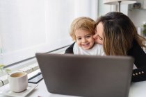 Femme employée à distance tenant un curieux petit enfant à genoux assis à table et travaillant avec un ordinateur portable à la maison — Photo de stock